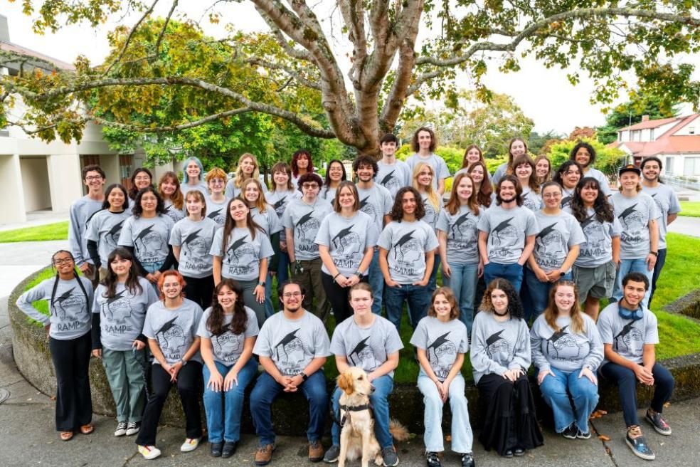 A large group of students sitting in front of a tree smiling, wearing grey matching shirts. 
