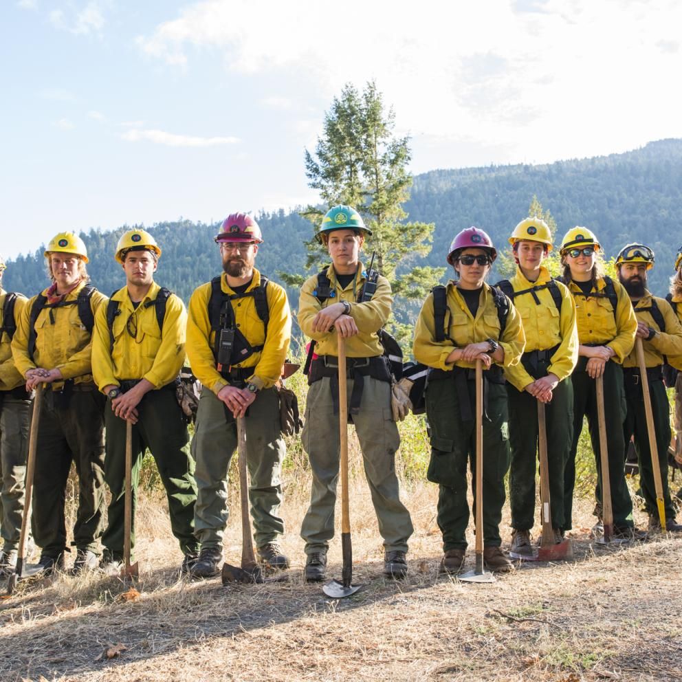 a line of students in fire gear standing in an open field with mountains in the background