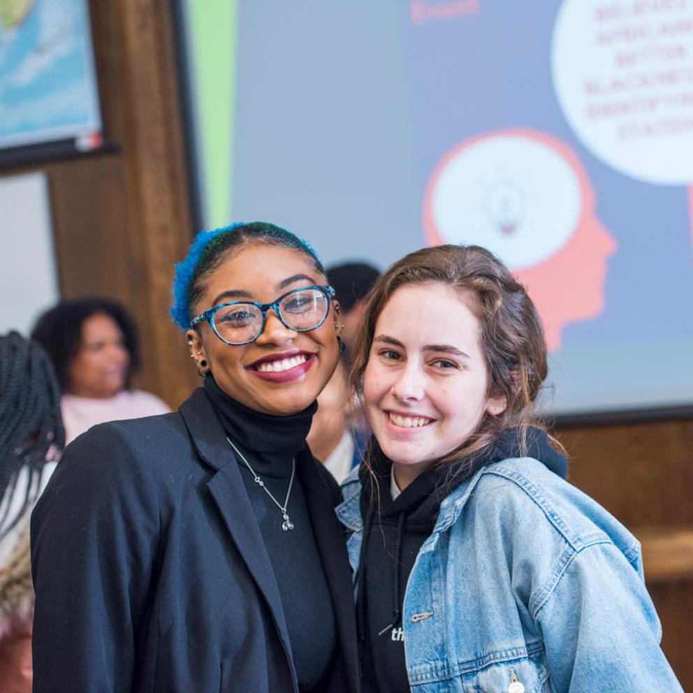 two students smiling at the camera with a presentation behind them