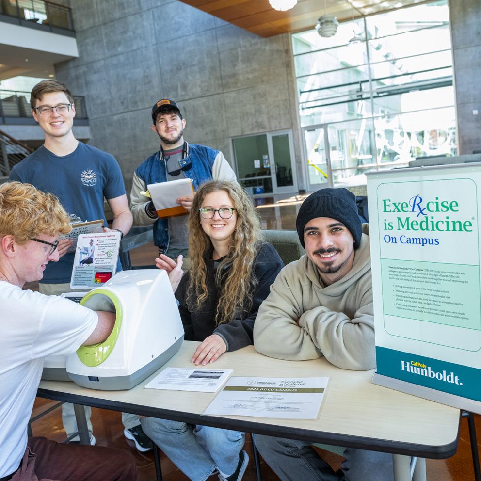 Students sitting at a table with one person putting his arm into a measuring maching