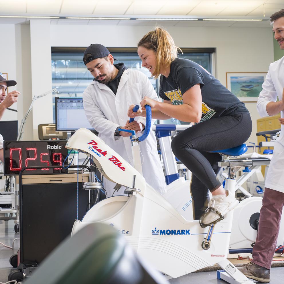 Student on a stationary bicycle being recorded by three other students in lab coats
