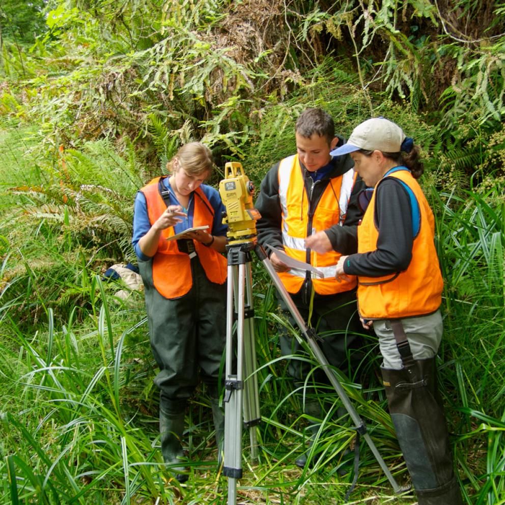 three students standing around a tripod with equipment on it and surrounded by green vegitation