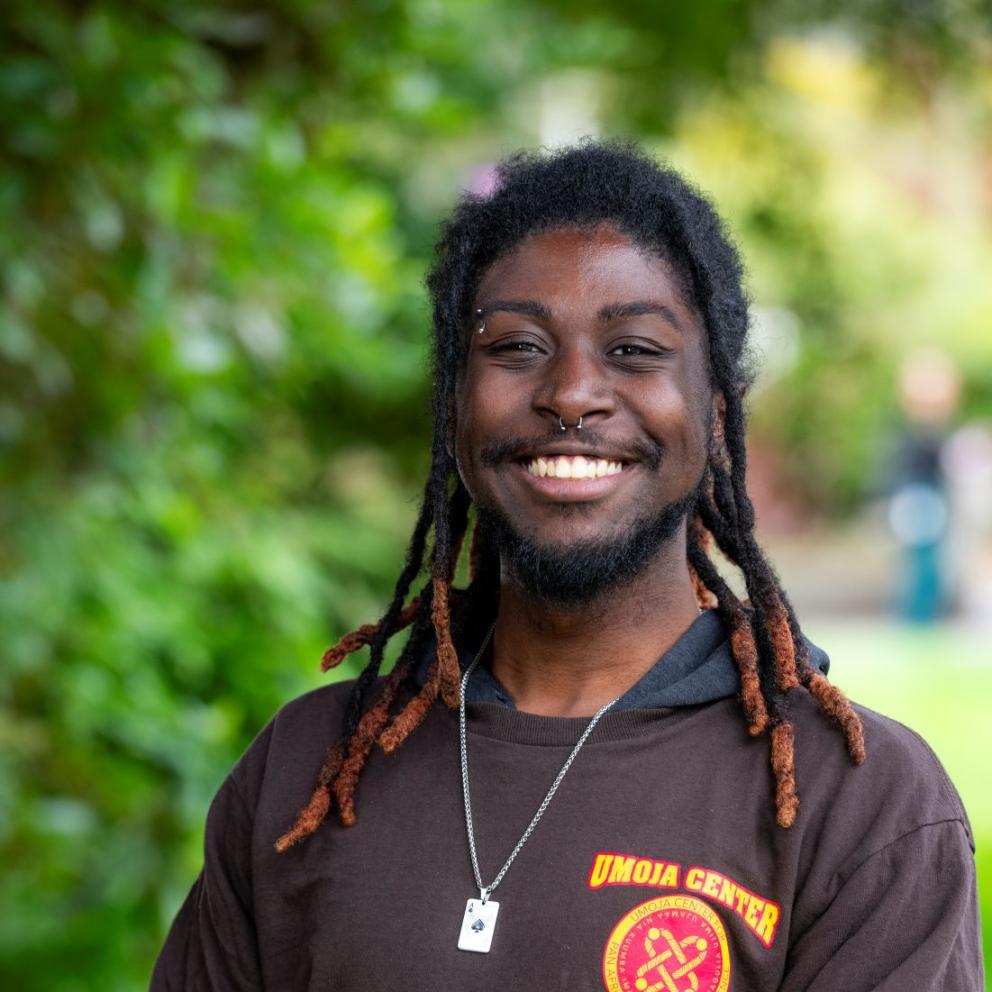 Kamar Little smiles while wearing their staff shirt which features the Umoja logo on a long sleeve chocolate brown shirt.