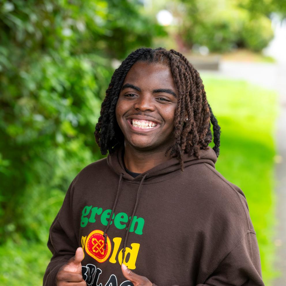 Shawn smiles while wearing their staff shirt which features the Umoja logo on a long sleeve chocolate brown sweatshirt.