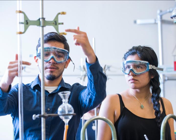 Two students with goggles on using chemistry equipment