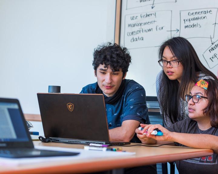 three students sitting at a table in front of a laptop