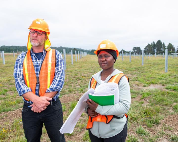 two people wearing hard hats carrying a clipboard and papers out in the field