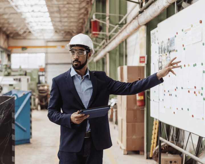 Man with hard hat standing in a warehouse pointing at a whiteboard