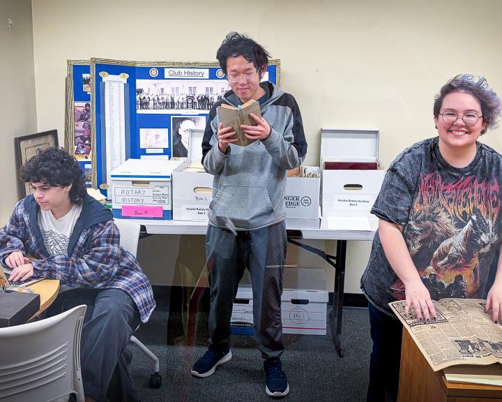 three students working on a project near a newspaper, typing on laptop and reading a book