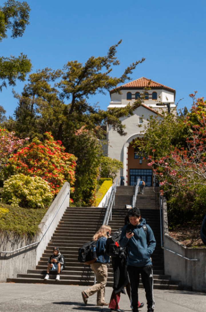 Students walk near the stairs leading up to Founders Hall, surrounded by colorful trees and plants on a sunny day.