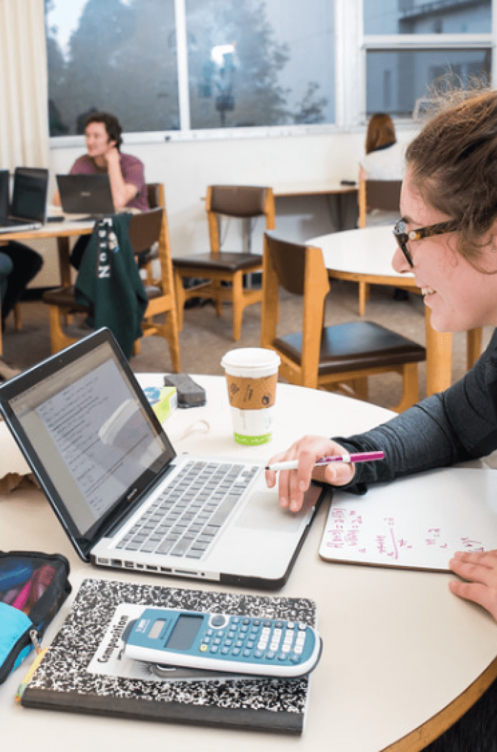 Student working on a laptop and whiteboard at a study table, with others collaborating in the background.