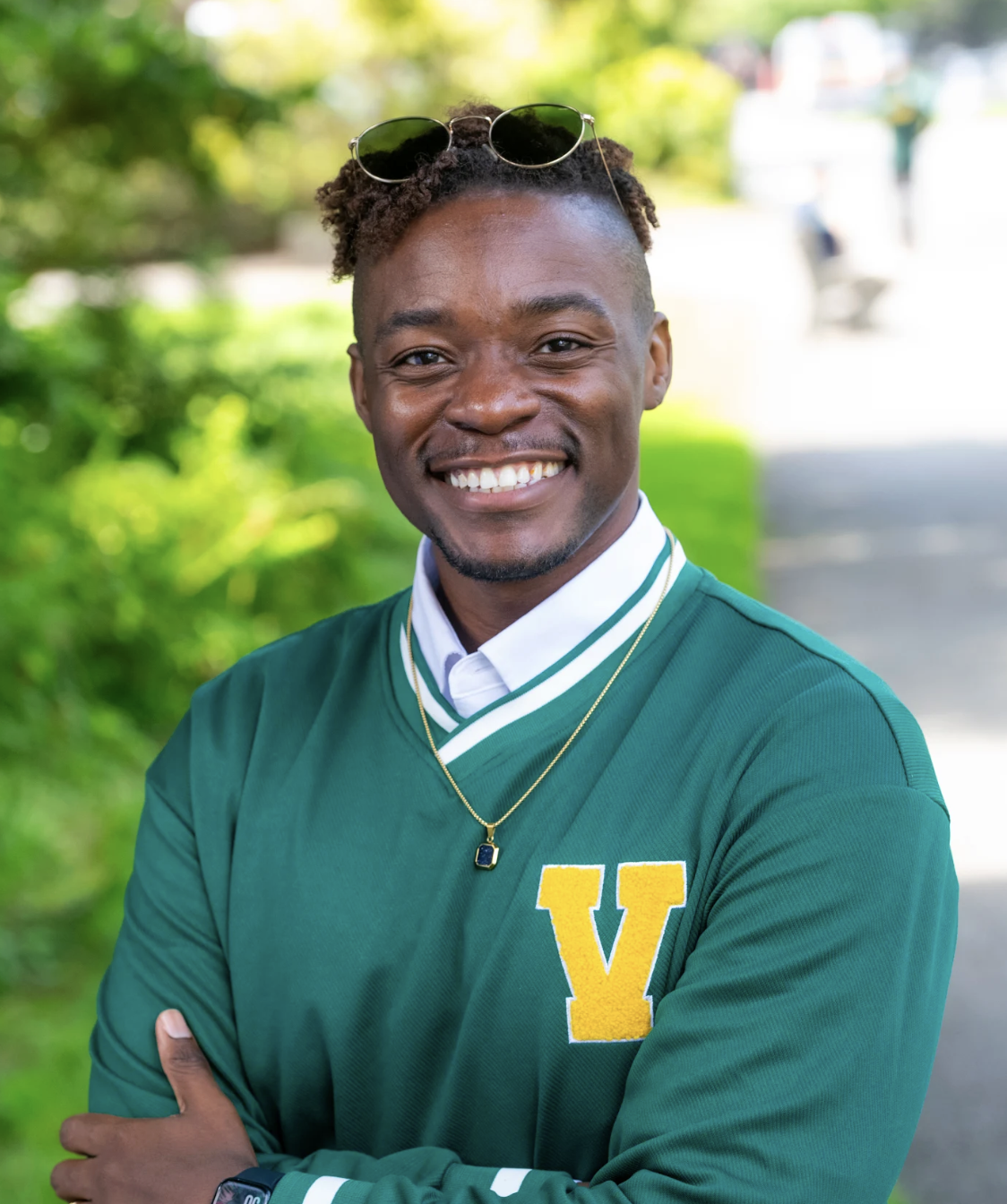 Travis Richards is smiling against a natural background. He has short hair cut with a bald fade and is wearing a green varsity crewneck.